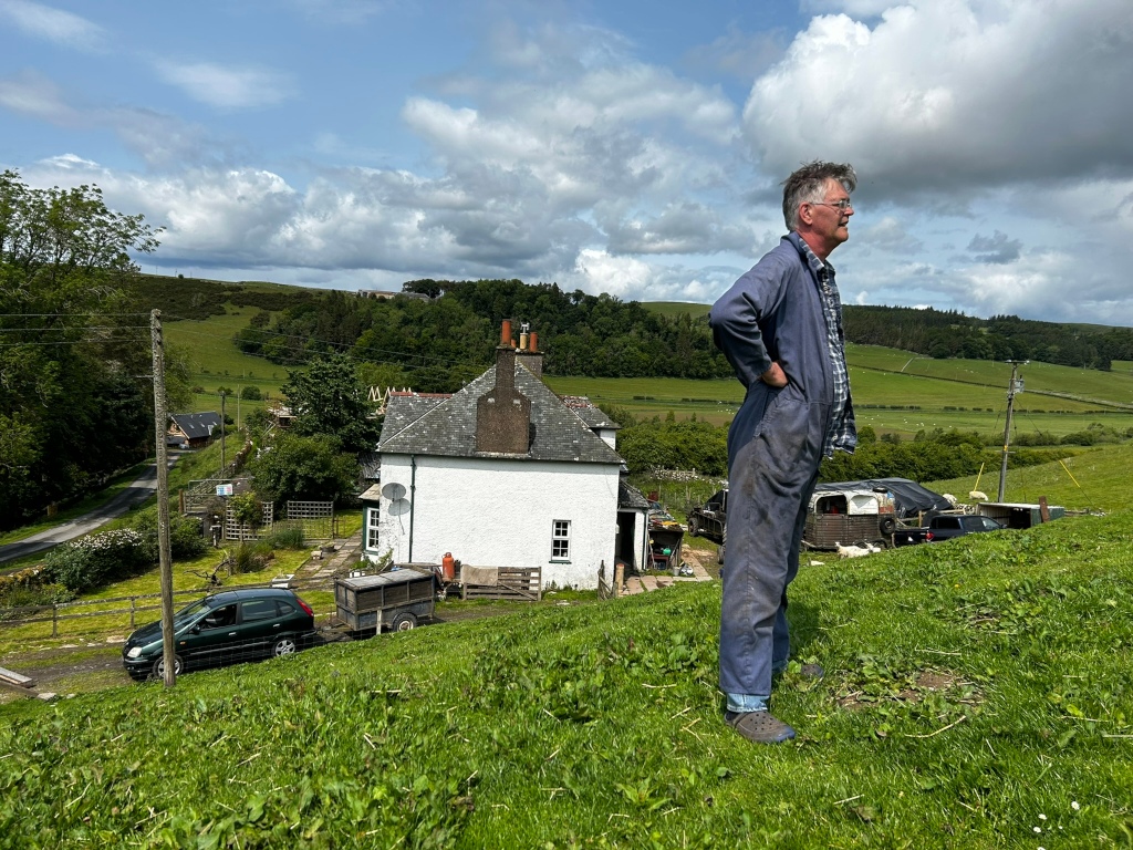 Charlie in work overalls standing in a grassy field, looking towards a white farmhouse in the background. The setting appears rural, with rolling green hills and scattered sheep visible in the distance. A dark-coloured car and a trailer are parked near the house. The sky is partly cloudy. The overall impression is one of rural tranquillity and possibly a sense of contemplation or observation by Charlie.