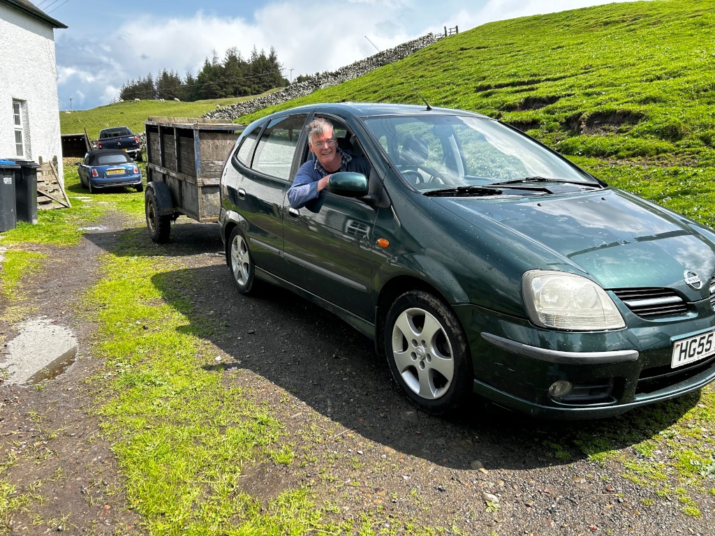 Charlie smiling whilat sitting in the driver's seat of a dark green Nissan Almeria. He's parked on a gravel driveway in front of a stone building in a rural setting with green hills in the background. The car is towing a small, wooden trailer. Another vehicle is parked further up the driveway.