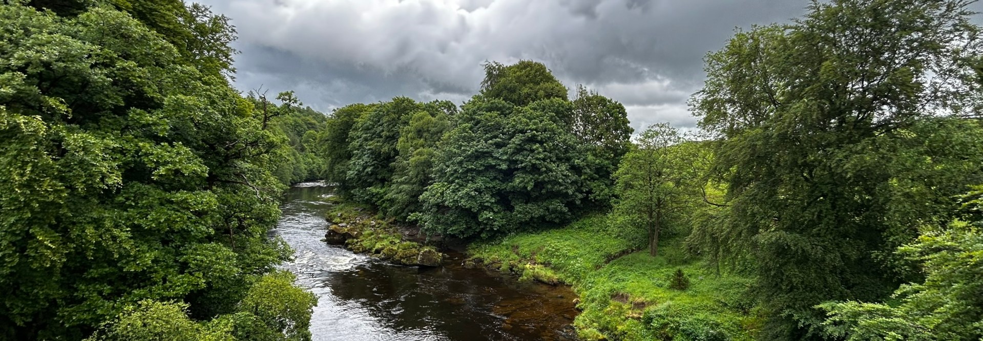 River flowing through a lush green valley. The river is relatively calm, with some gentle ripples visible. The banks are densely covered with trees and vegetation, creating a feeling of seclusion and tranquillity. The sky is overcast, adding to the serene and somewhat moody atmosphere.