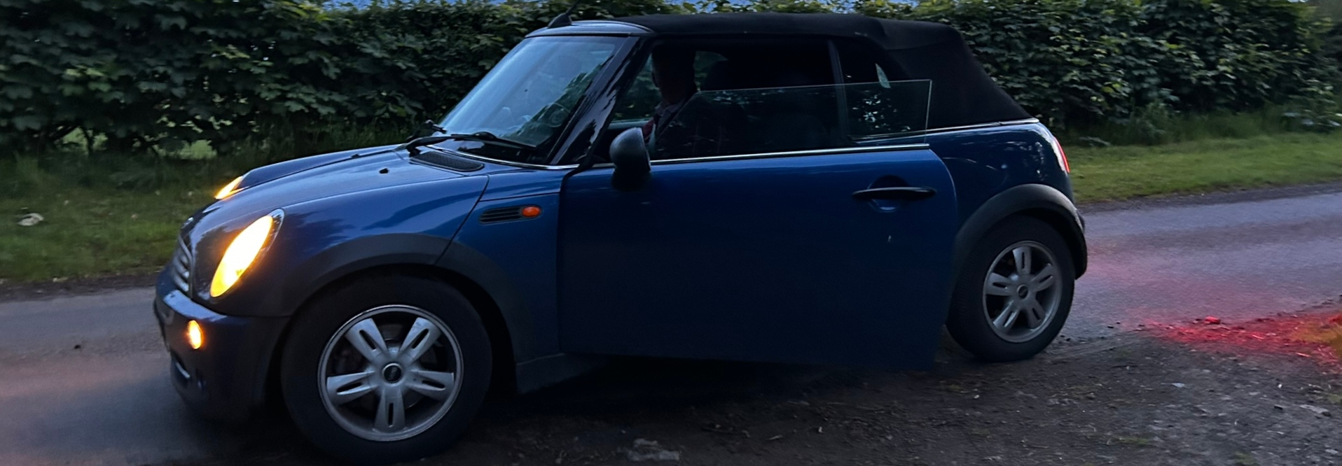 Panoramic view of a vast, gently rolling field of tall, green barley swaying in the breeze under a dramatic sky.  A small copse of trees sits atop a rise in the middle ground. The sky is a mix of soft blues and darker, heavier clouds suggesting an approaching storm or the aftermath of one. A crescent moon is visible in the upper left corner, adding to the serene yet somewhat ominous atmosphere. The overall feeling is one of peaceful vastness juxtaposed with the impending change hinted at in the sky.