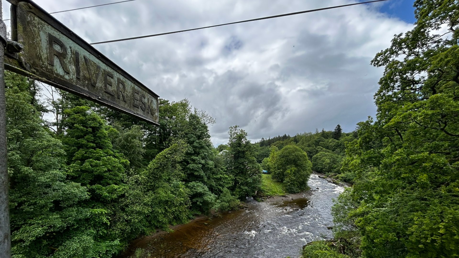 Slightly overcast day view of the River Esk in Scotland, as indicated by a weathered sign. The river flows through a lush green valley, bordered by dense trees and vegetation. The water appears somewhat turbulent, suggesting a gentle current. The overall mood is peaceful and serene, showcasing the natural beauty of the Scottish countryside.