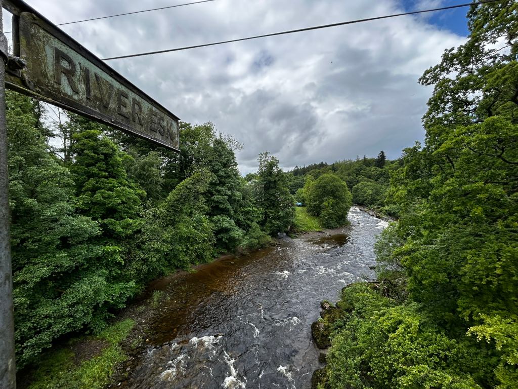 Slightly overcast day view of the River Esk in Scotland, as indicated by a weathered sign. The river flows through a lush green valley, bordered by dense trees and vegetation. The water appears somewhat turbulent, suggesting a gentle current. The overall mood is peaceful and serene, showcasing the natural beauty of the Scottish countryside.