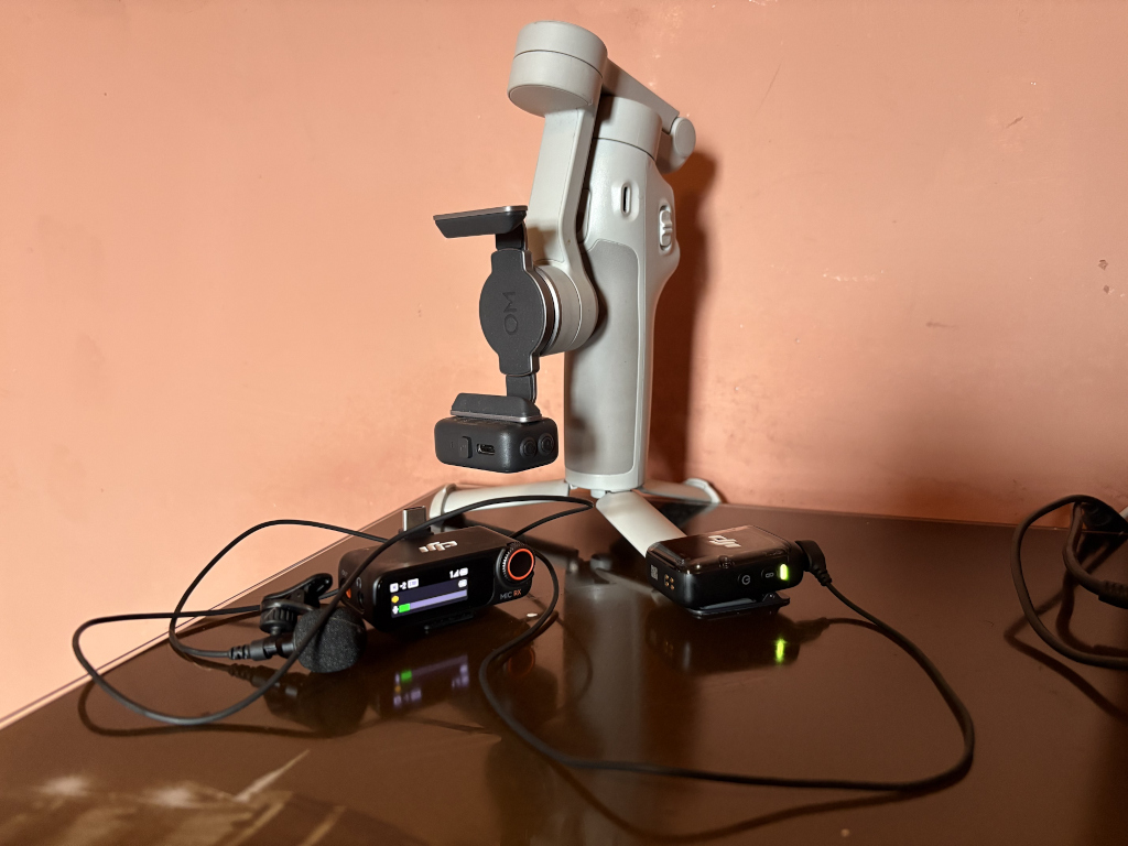 DJI OM 7 smartphone gimbal, mounted on a tripod, with wireless microphones placed on a dark surface below it. The microphones are connected via cables, indicating they are ready for use in a video recording setup. The overall composition suggests preparation for filming or content creation.