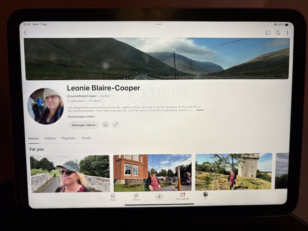 iPad screen displaying the YouTube channel of Leonie Blaire-Cooper. The screen is slightly angled, and the top edge is partially cut off. The central focus is Leonie's YouTube profile. Her profile picture shows a headshot of a woman with shoulder-length brown hair, wearing a cream-colored beanie and a dark jacket. She appears friendly and approachable. Below the profile picture is her channel name in a bold, slightly stylised font, followed by her bio, subscriber count, and video count. Several thumbnails of her videos are displayed below, showcasing picturesque Scottish landscapes, including a stone tower, a quaint village setting, and a woman (presumably Leonie) walking through a field.