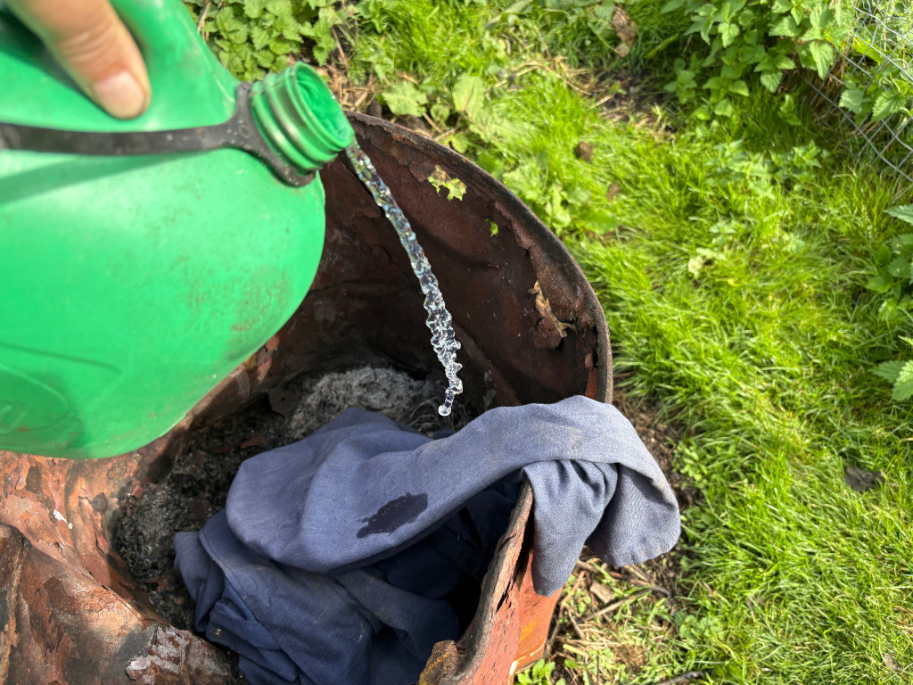 Leonie's hand pouring water from a green plastic jug into a rusty metal barrel.  Inside the barrel, a dark blue piece of clothing is visible. The barrel is situated outdoors on the ground, surrounded by grass and some weeds. The scene suggests a simple, possibly outdoor, cleaning or disposal activity.