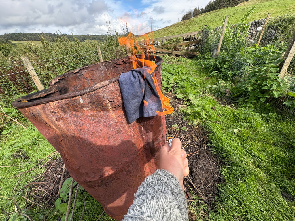 Eye-level view of a Leonie's hand holding a lighter near a rusty, metal barrel. A large, rusty metal barrel, seemingly an old oil drum, is the central focus. It's significantly rusted and shows signs of age and weathering. A piece of dark-coloured fabric is burning inside it, creating flames that are visible at the top. Leonie's  hand, wearing a light grey fuzzy sweater, is holding a lighter near the barrel, suggesting she is are either lighting or maintaining the fire.