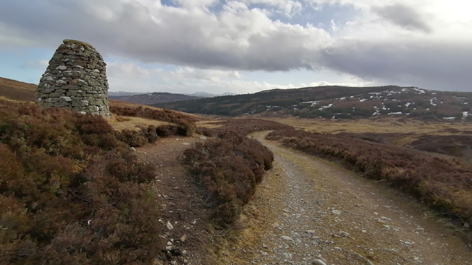 A serene, misty mountain landscape. A calm, still lake sits nestled within a valley, surrounded by low-lying, brown and green vegetation. The mountains rise steeply on either side, their peaks shrouded in low-hanging clouds or mist. The overall mood is peaceful and somewhat melancholic, due to the muted colors and atmospheric conditions. There's a sense of solitude and the vastness of the natural world.