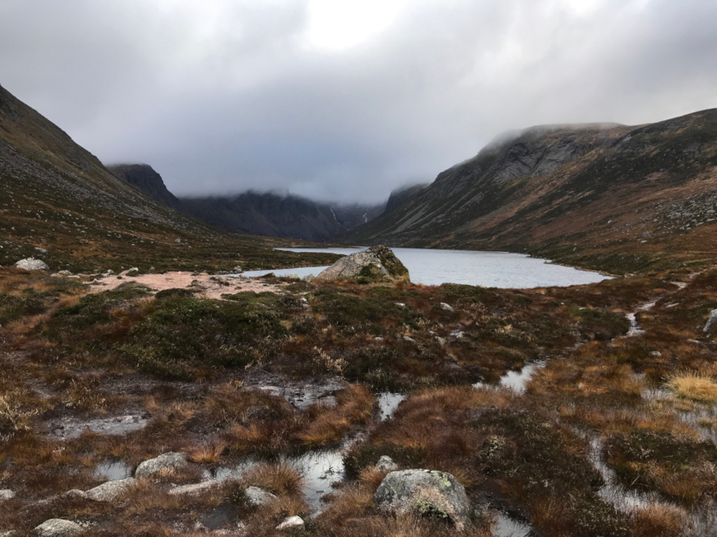 Serene, misty mountain landscape. A calm, still lake sits nestled within a valley, surrounded by low-lying, brown and green vegetation. The mountains rise steeply on either side, their peaks shrouded in low-hanging clouds or mist. The overall mood is peaceful and somewhat melancholic, due to the muted colours and atmospheric conditions. There's a sense of solitude and the vastness of the natural world.