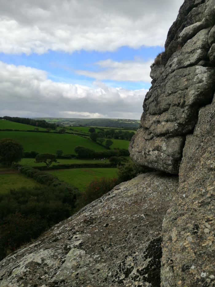 Rocky outcrop, possibly a tor, in the foreground. The rock is grey and textured, appearing weathered. The background displays a lush green pastoral landscape of rolling hills and fields, under a partly cloudy sky with patches of blue. A few trees and what appear to be farm animals can be seen in the distance. The overall impression is one of a serene, rural scene viewed from an elevated vantage point.