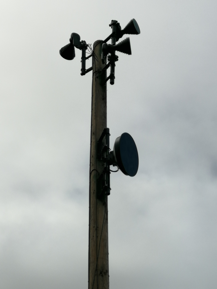 Wooden utility pole with various pieces of equipment attached. Near the top are several dark, horn-shaped devices, possibly lights or speakers. Lower down is a dark, circular dish-shaped antenna or similar device. The sky is overcast and grey.