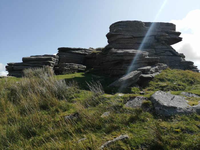 Rocky outcrop on a grassy hillside, possibly in a moorland or upland area. The rocks are layered and appear to be weathered, suggesting a natural formation. The grass is short and somewhat windswept. Sunlight creates a lens flare effect in the upper right portion of the image.