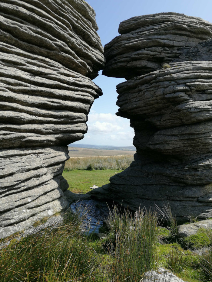 Natural rock formation, appearing as two large, layered rock faces that meet, creating a narrow opening. Through this opening, a small pool of water is visible, leading to a grassy plain and a distant horizon under a clear blue sky. The rocks are grey and show clear layers of stratification.