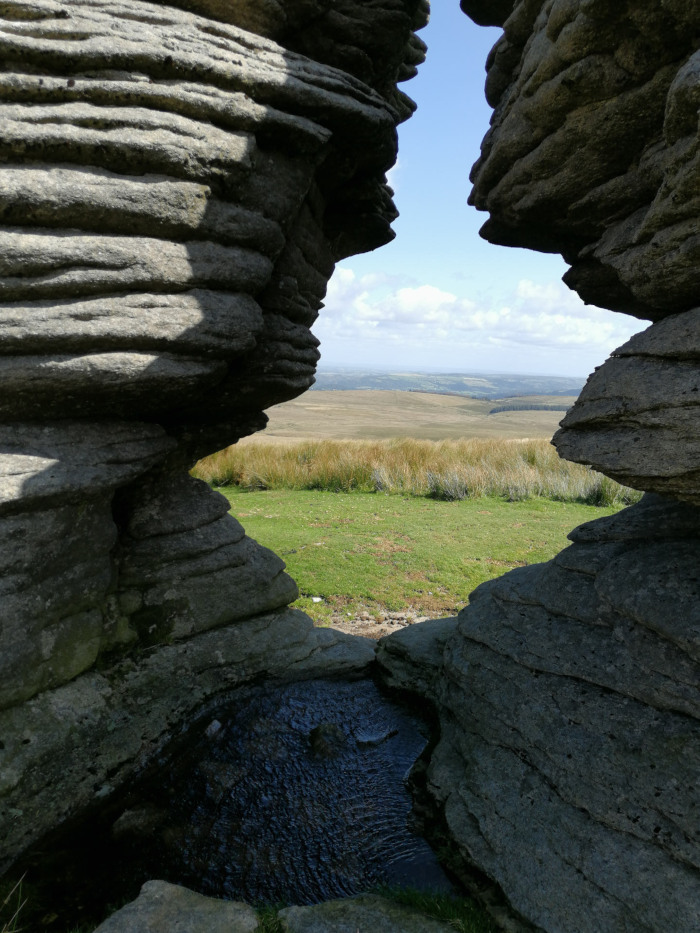 Natural rock formation creating a frame-like opening. Through this opening, a pastoral landscape of a green field and distant hills under a partly cloudy sky is visible. A small pool of water sits at the bottom of the rock formation within the frame. The layered texture of the rocks is prominent.