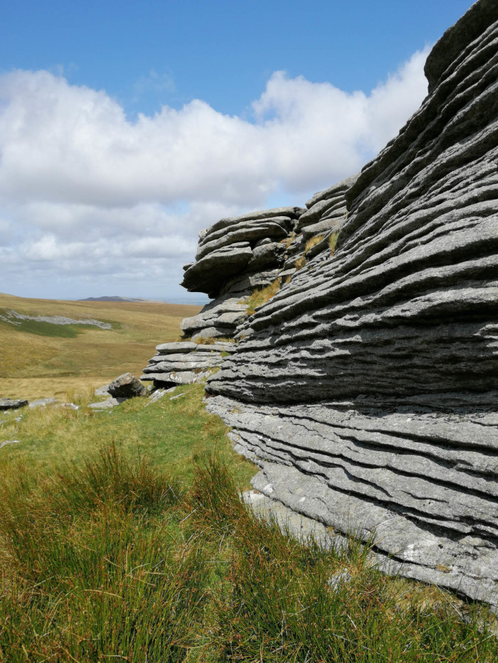 Natural rock formation creating a frame around a view of a grassy landscape. The rocks are layered and appear weathered, forming a kind of window or opening. A small pool of water sits at the base of the rocks within this frame. Beyond, a pastoral scene of rolling hills and fields under a partly cloudy sky is visible.