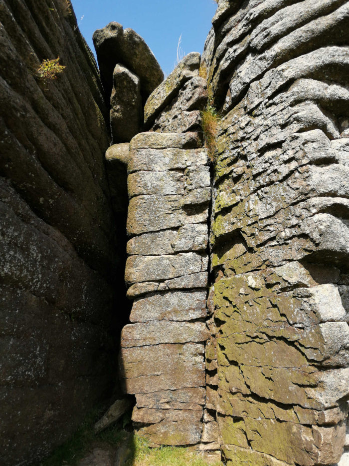 Close-up view of a rock formation. Two large sections of layered rock meet at a narrow crevice, creating a vertical fissure. The rocks are predominantly brown and gray, showing signs of weathering and erosion. Patches of green lichen or moss are visible on some surfaces. The texture is rough and uneven.