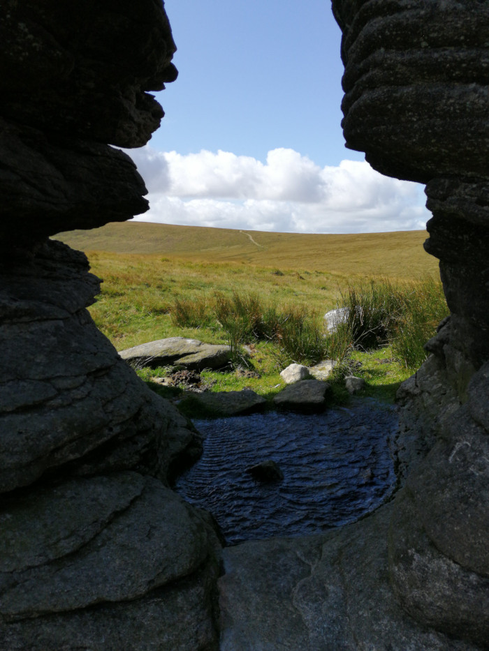 Natural rock formation creating a frame around a view of a moorland landscape. A small pool of still water sits at the base of the rocks in the foreground, while the background features rolling hills under a partly cloudy sky.
