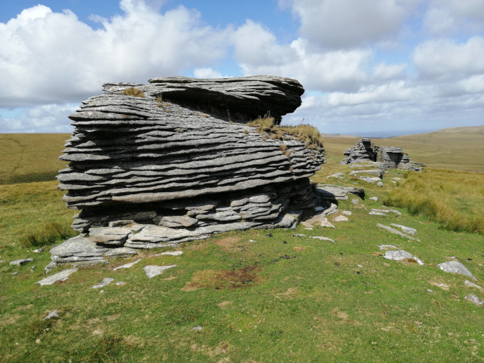 Landscape dominated by a large, layered rock formation in the foreground. The rock is grey and appears to be sedimentary, with distinct horizontal strata. It sits on a grassy hillside under a partly cloudy sky. In the background, another, smaller rock formation is visible, along with a relatively flat, grassy expanse extending to the horizon.