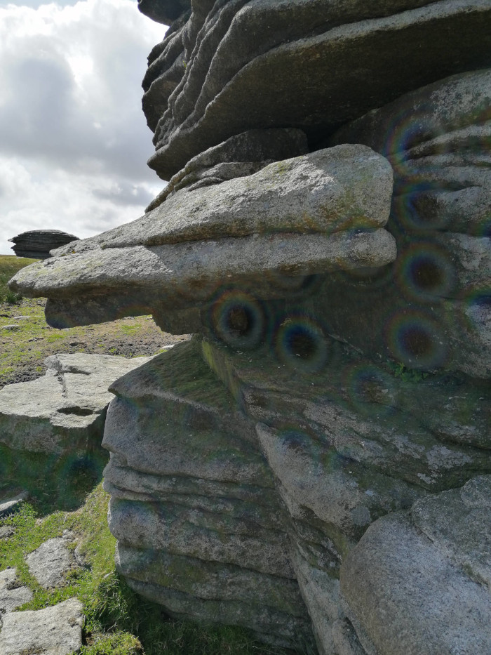 Close-up view of a rocky outcrop, possibly in a moorland or upland area. The rocks are layered and appear weathered, with a variety of textures and shades of grey. There's some green vegetation visible at the base of the rocks. The overall impression is one of rugged, natural beauty and the passage of time. Lenses flares are visible upon the rocks surface.