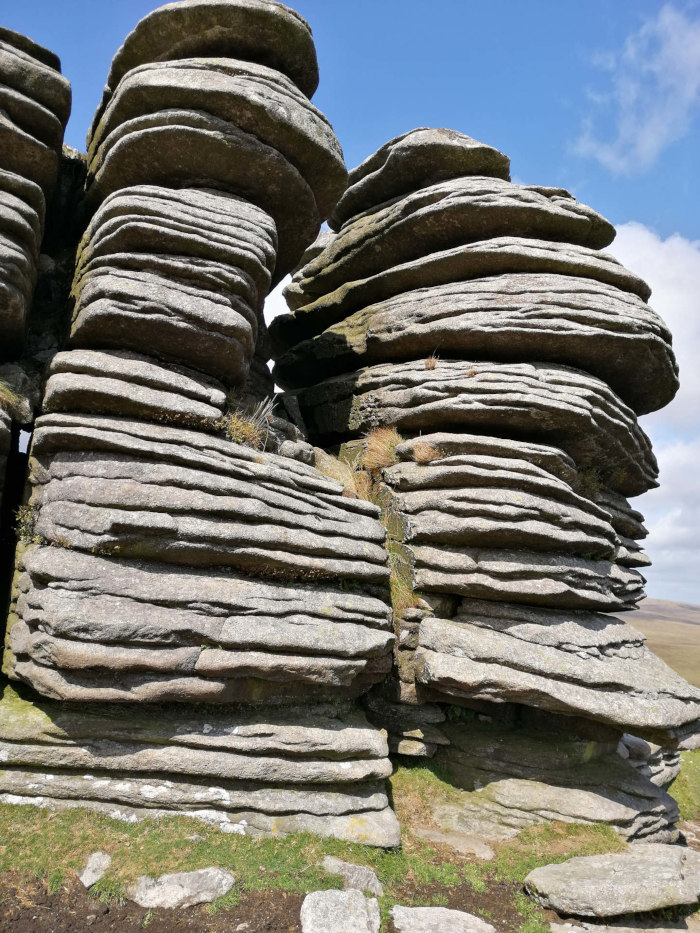Close-up view of a rock formation. The rock is layered, appearing as a stack of large, flat stones, with a natural fissure running between two sections of the formation. Sparse vegetation is visible in crevices between some of the rock layers. The background shows a clear blue sky and a hint of distant landscape. The overall impression is one of rugged, natural beauty.