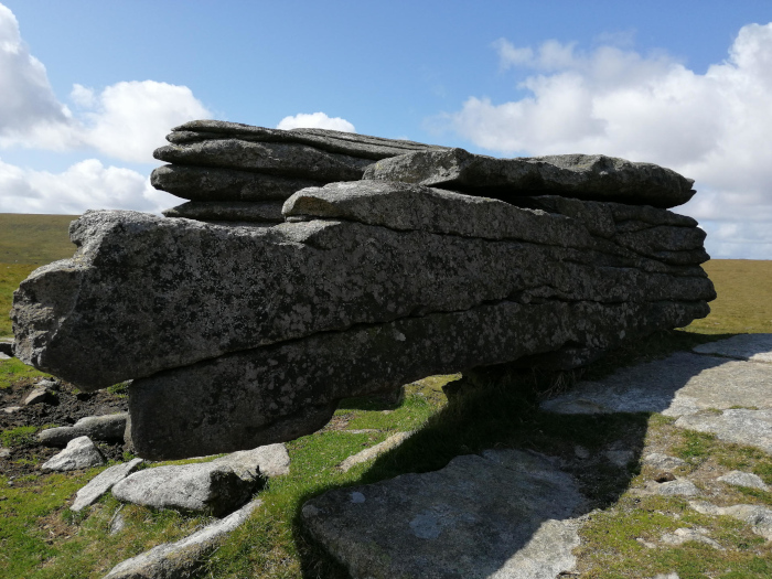 Large, layered rock formation, seemingly weathered and eroded over time. It's situated in a grassy landscape under a partly cloudy sky. The rock's horizontal strata are clearly visible, suggesting geological processes like sedimentation and uplift. The overall impression is one of natural beauty and the power of natural forces shaping the landscape.