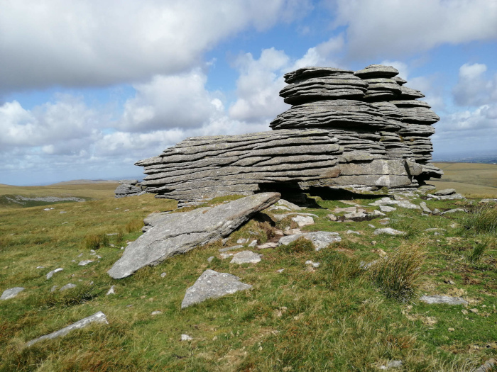 Large, layered rock formation situated in a grassy field under a partly cloudy sky. The rock is the central focus, exhibiting a stratified structure with numerous horizontal layers. The surrounding landscape is relatively flat and sparsely vegetated.