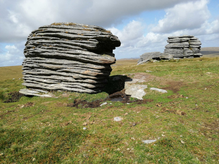 Two large rock formations, layered and appearing almost man-made in their precise, stacked structure. They sit in a grassy field under mostly cloudy skies. The larger rock is in the foreground, partially obscuring a small puddle of collected water.