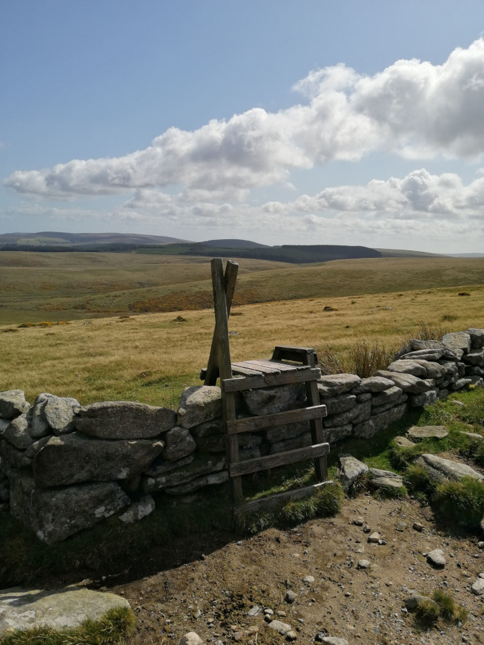 Rustic wooden stile built into a low stone wall, set against a backdrop of expansive, rolling moorland under a partly cloudy sky. The scene is peaceful and evokes a sense of quiet, rural solitude. The stile acts as a visual focal point, suggesting a pathway through the landscape and inviting the viewer to explore the vast open space beyond.