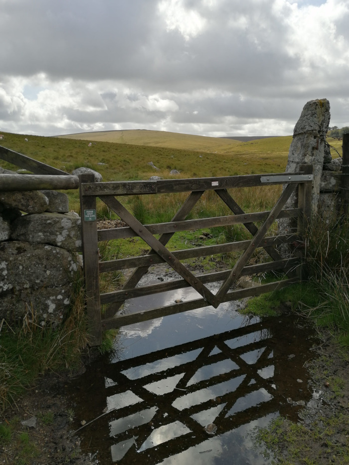 Wooden gate set in a stone wall, spanning a small, muddy stream or puddle. Beyond the gate stretches a vast, open moorland under a cloudy sky. The gate's reflection is clearly visible in the water. Sheep are visible in the distance on the moorland. A small sign on the gate post requests that people close the gate.