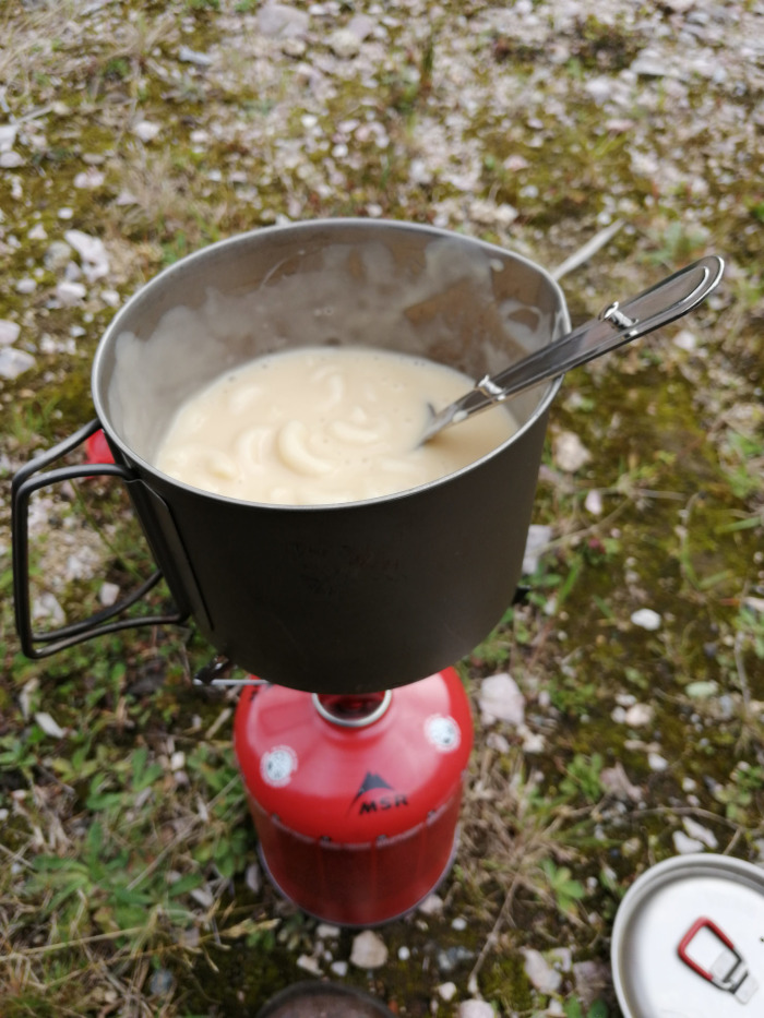Pot of creamy, light-coloured food, possibly a soup or stew, cooking on a portable camping stove outdoors. A spoon rests in the pot. The setting appears to be a grassy, somewhat rocky area.