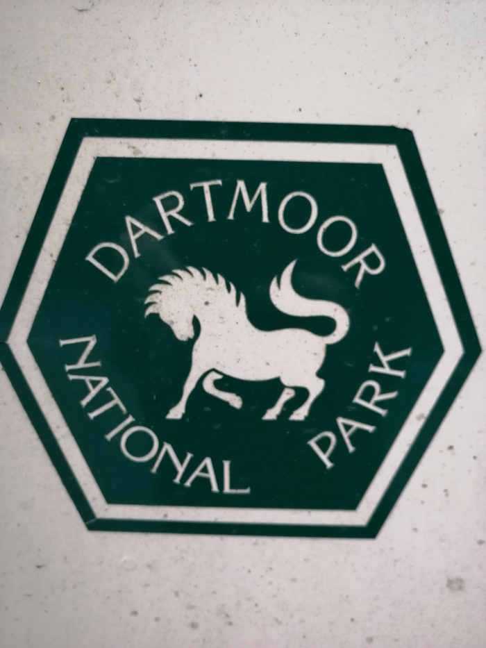 Dark-green hexagonal sticker or sign. The text Dartmoor National Park is written around the circumference, with a stylised white image of a pony in the centre.