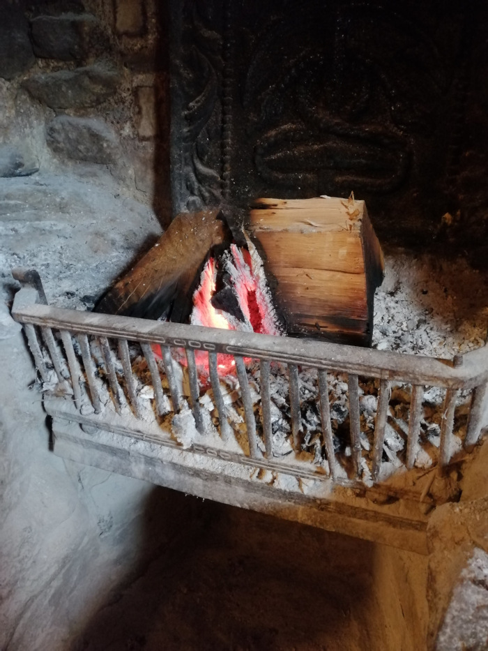 Close-up view of a fireplace with a fire burning inside. A metal grate holds the burning wood and ash. The fireplace's interior is made of stone, and there's a decorative, dark, possibly metal, back plate visible behind the fire. The overall impression is one of rustic warmth and age, suggesting a traditional or older home setting.
