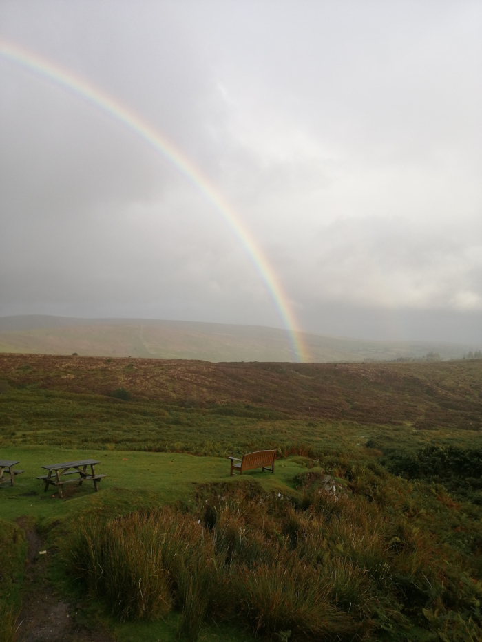 Scenic landscape featuring a partially visible rainbow arching over a gently rolling, grassy hill. In the foreground, there's a wooden bench and a picnic table, suggesting a resting place for hikers or visitors. The overall mood is serene and peaceful, enhanced by the post-rain atmosphere. The horizon displays a vista of rolling hills, extending into the distance.