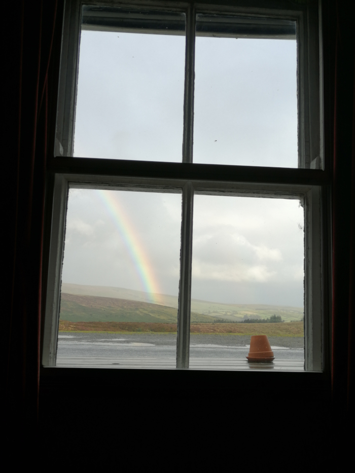 View from an indoor window.  A vibrant rainbow arcs across a landscape of rolling hills visible through four panes of glass. A small terracotta pot sits on the windowsill in the foreground. The overall mood is serene and peaceful, with a sense of calm after a rain shower.