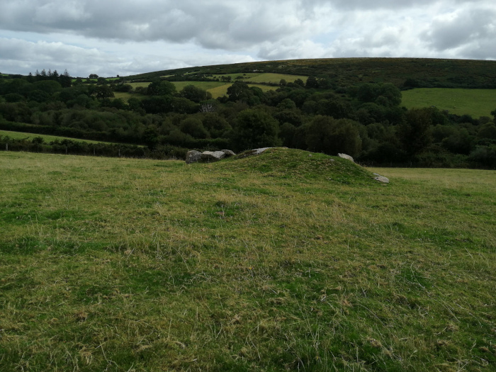 Grassy field in a rural setting. In the foreground is a low, grassy mound with a few large stones visible on its top and sides. The background features a verdant landscape of rolling hills, fields, and trees under a somewhat cloudy sky.
