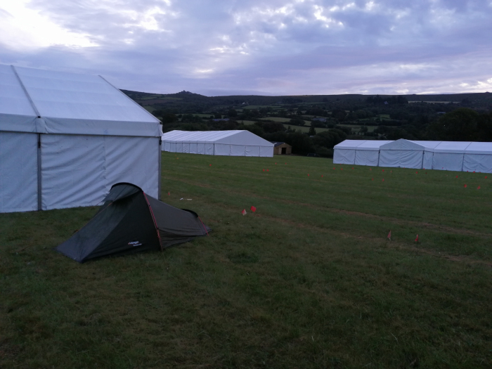 Grassy field with several large white tents in the background. In the foreground, a small, dark-coloured tent is pitched close to one of the larger tents. The background includes a gently rolling landscape under a cloudy sky; a hill or small mountain is visible in the distance. The overall impression is of a temporary camp or festival site.