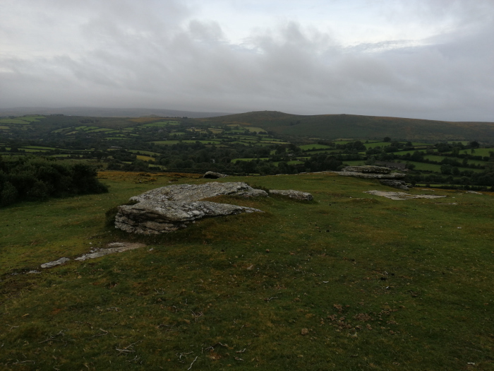 Picturesque landscape on an overcast day. In the foreground, there are some weathered, light-grey rocks sitting on a short, green grassy hill. The midground and background display a rolling, verdant valley, dotted with patches of trees and farmland under a cloudy sky.