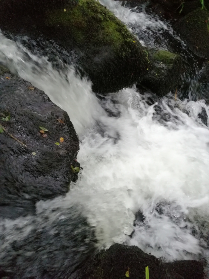 Close-up view of a small waterfall or cascade. Water rushes over dark, moss-covered rocks, creating white frothy water. The focus is on the turbulent movement of the water as it flows over the rocks.