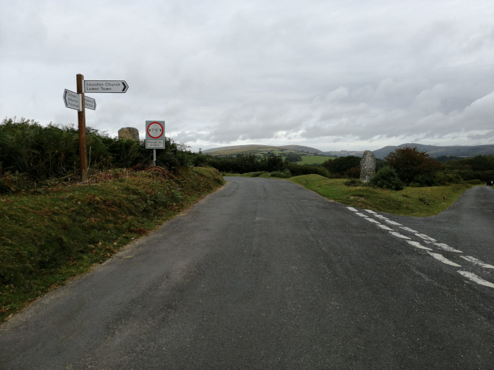 Rural road fork in the countryside under a cloudy sky. A wooden signpost indicates directions to Leusdon Church and Lower Town. A no-overtaking sign is also visible. In the background, rolling hills and a standing stone are present.