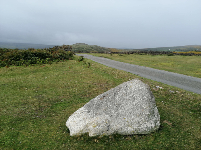 Large, light gray boulder resting in a grassy field beside a paved road. In the background, rolling hills and low vegetation extend to the horizon under a muted, overcast sky. The overall impression is one of a serene, somewhat desolate, rural landscape.