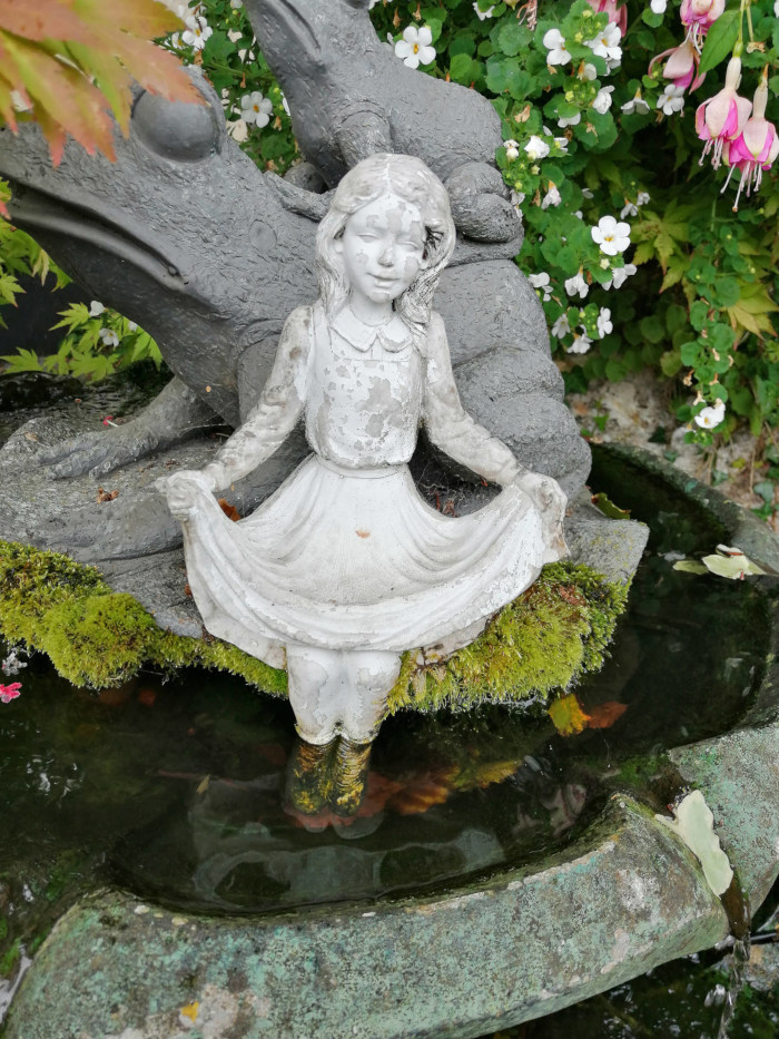 Garden fountain. A small, weathered, stone statue of a girl in a dress is positioned in the center of the fountain's basin. The girl statue is seated, and her dress appears to be billowing slightly. Behind the girl statue are two larger, gray stone statues of what seem to be rabbits or hares. The fountain basin is itself made of stone, showing signs of age and moss growth along its edges. The fountain is surrounded by lush greenery, including flowering plants.