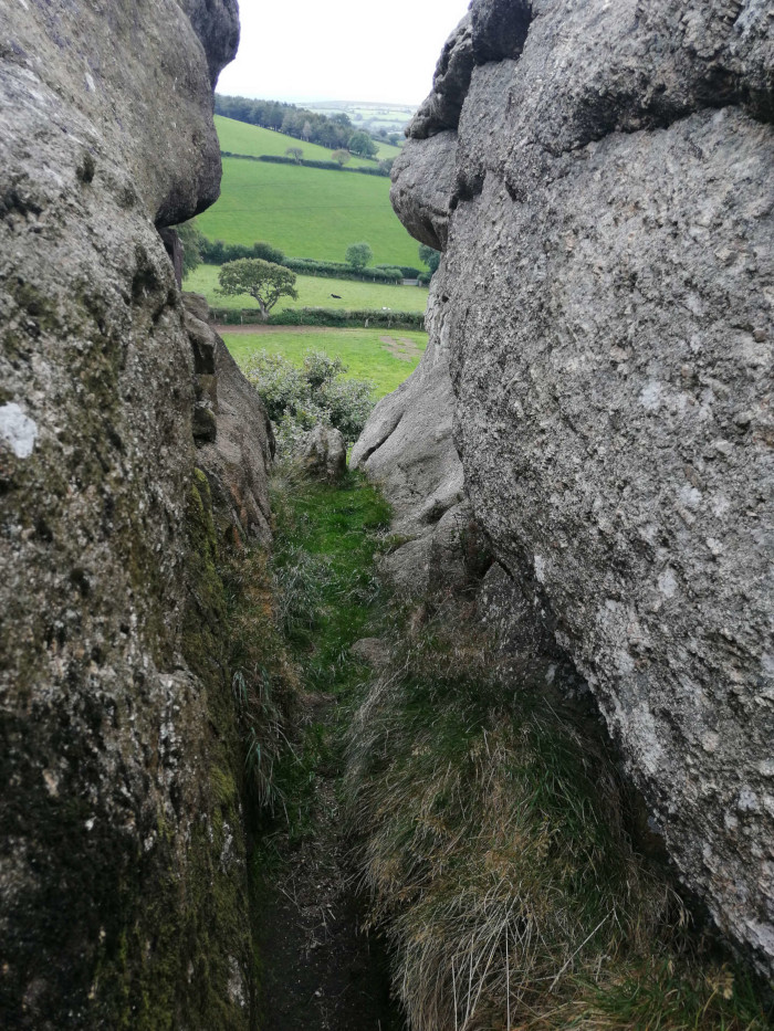 Narrow passage between two large, grey rocks.  Green grass and vegetation grow in the crevice and beyond, leading the eye to a view of a verdant, rolling hillside in the distance. The overall impression is one of a secluded natural pathway offering a glimpse of open countryside.