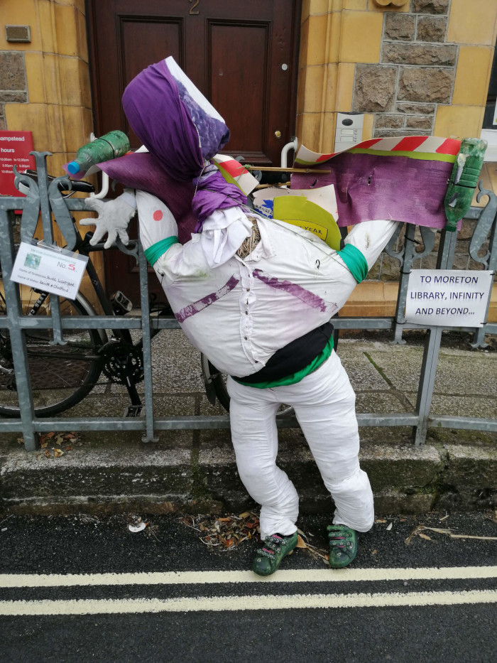 Scarecrow, creatively constructed and seemingly participating in a scarecrow festival or community event. The scarecrow is designed in a humorous yet slightly chaotic manner. It’s leaning against a metal fence, next to a bicycle, and appears to be dynamic rather than static. It is wearing what looks like repurposed clothing and has various materials attached: a purple cloth as a head covering, partially visible posters and other discarded items. It has shoes on its feet, adding to its intentionally quirky appearance.
