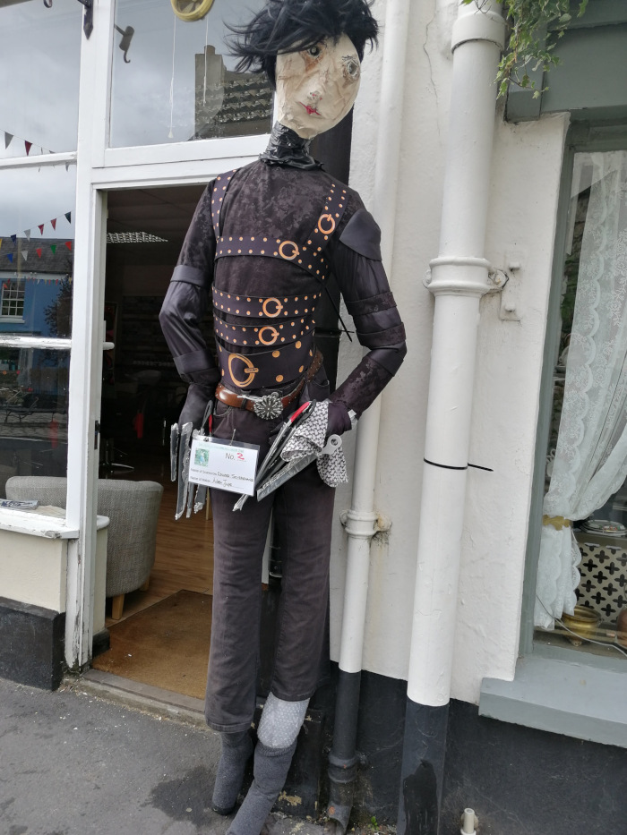 Scarecrow dressed as Edward Scissor hands, the character from Tim Burton's movie. The scarecrow is positioned outside a shop, leaning against a wall. It's meticulously crafted to resemble the character, including the dark clothing, scissor hands, and dark hair. A small identifying placard is visible on its waist.