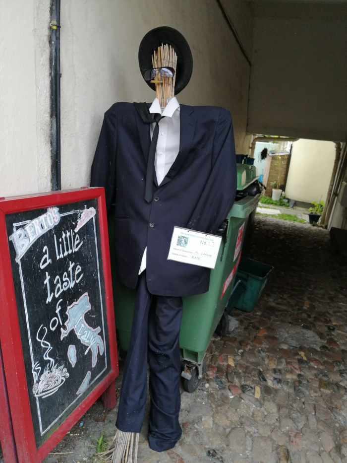 Scarecrow dressed in a dark suit, sunglasses, and a fedora hat. The scarecrow is positioned next to a red sign advertising a little taste of Italy, suggesting a connection to an Italian restaurant. The background depicts a narrow alleyway. The overall impression is a humorous and creative display, possibly part of a scarecrow festival or similar event.