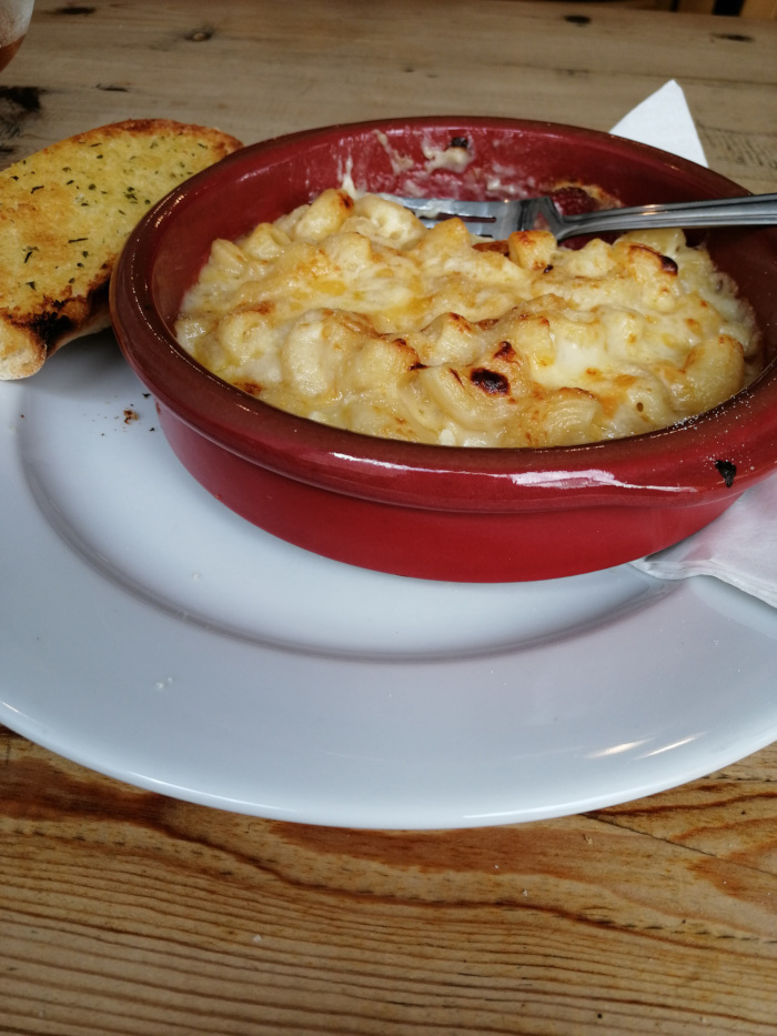 Small, red, ceramic dish of macaroni and cheese. A piece of garlic bread is visible on a white plate next to the mac and cheese. The setting appears to be a casual dining table with a wooden surface. A fork is partially visible in the dish.