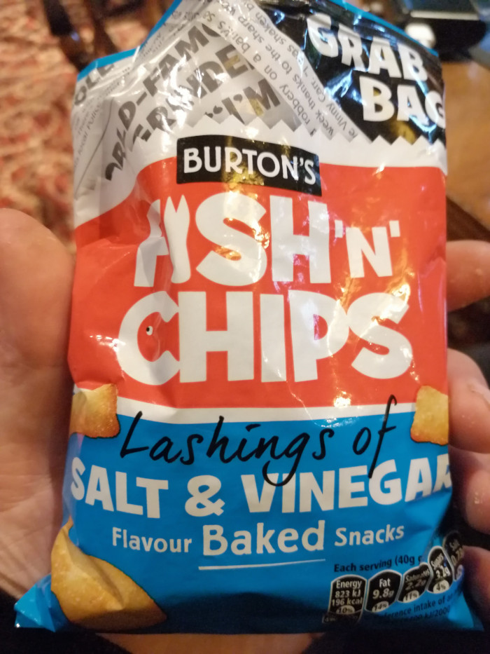 Leonie's hand holding a bag of Burton's Fish 'n' Chips, a baked snack with lashings of salt and vinegar flavour. The bag is predominantly red and blue, and features a graphic of a fish. The bag also advertises itself as a Grab Bag. The image is simple and focuses on the product itself. There's no deeper creative background, emotional expression, story or moral to analyse beyond the typical advertisement of a snack food.