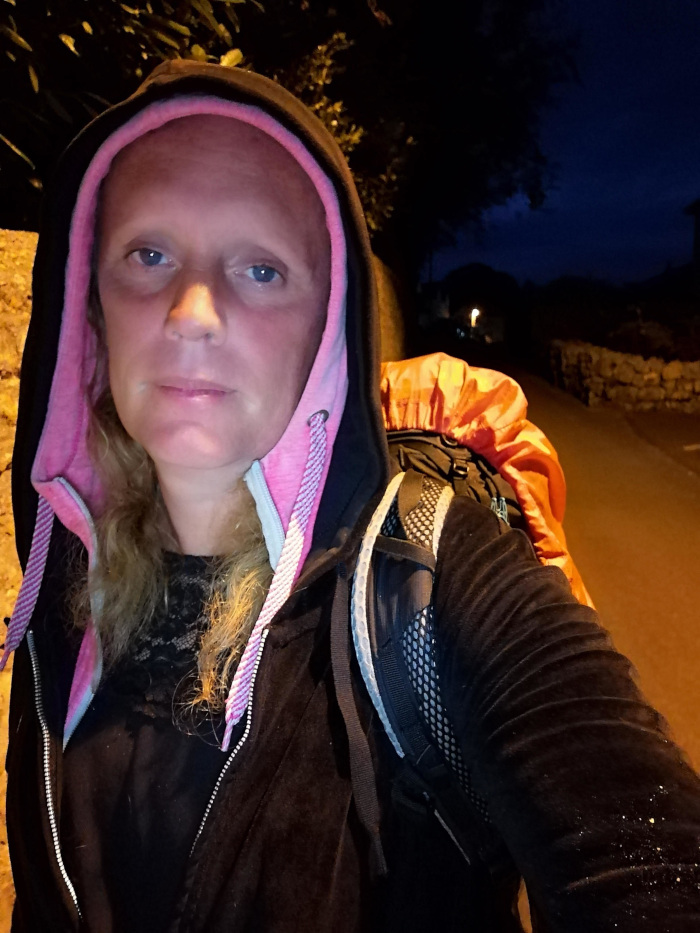 Leonie at night, standing on a street. She is wearing a black hoodie with a pink lining, a black jacket, and a large backpack. Her expression is serious or neutral. The background is dark, with a dimly lit street visible. The overall impression is one of nighttime travel or a journey.