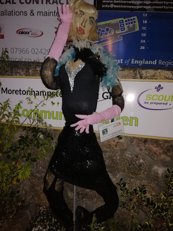 Scarecrow dressed as a glamorous woman. It's wearing a black sequined dress, pink gloves and sleeves, and a blonde wig. The scarecrow is posed with one hand raised to its face, and it's positioned against a stone wall with signage in the background that includes a business advertisement and a Scout group logo. The overall impression is one of whimsical humour and creativity. The scarecrow's attire suggests a celebration or festival, possibly a scarecrow competition given the number card visible next to it.