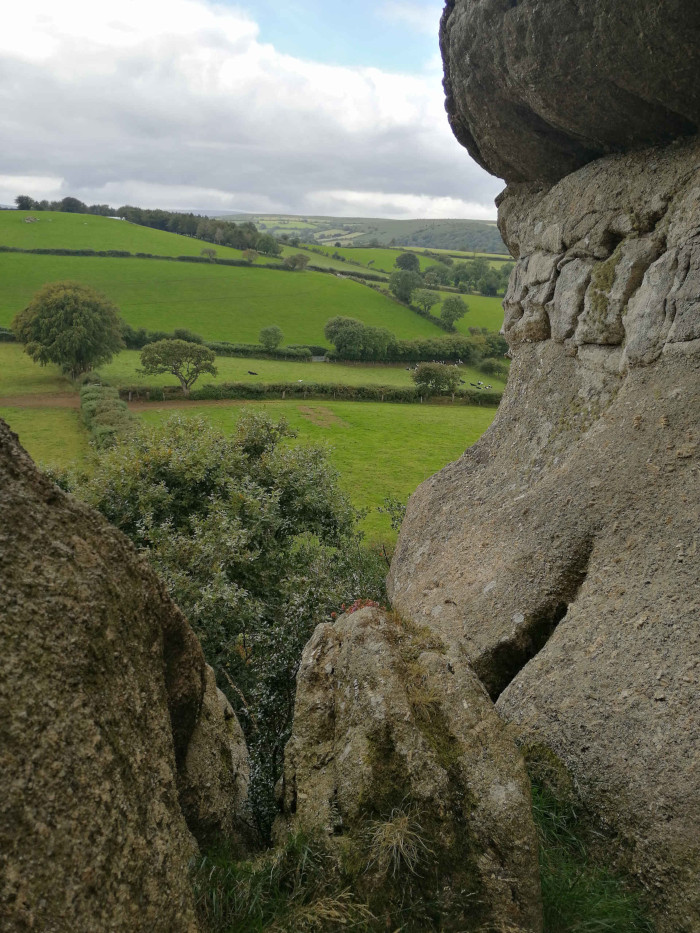 Verdant, rolling landscape of green pastures and hedgerows, typical of the British countryside. This pastoral scene is framed by large, grey, moss-covered rocks, creating a natural window-like view from a rocky outcrop or possibly a natural rock formation. The sky is slightly cloudy, providing soft, diffused light. The overall impression is one of peaceful tranquillity and the juxtaposition of rugged nature with calm farmland.