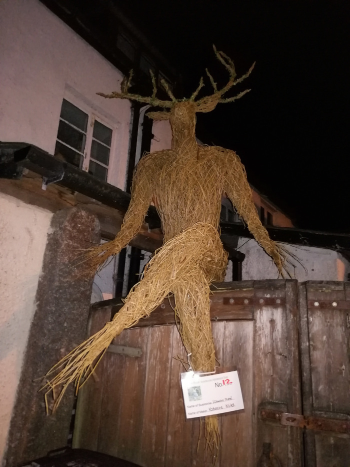 Whimsical, anthropomorphic sculpture of a deer crafted from straw or similar natural fibers. The figure is seated, with long, dangling legs, on a wooden gate or door at night in front of a building. A small label affixed to the sculpture provides identification details, suggesting it may be part of a larger exhibition or display, possibly a scarecrow contest. The overall impression is one of rustic charm and folk artistry.